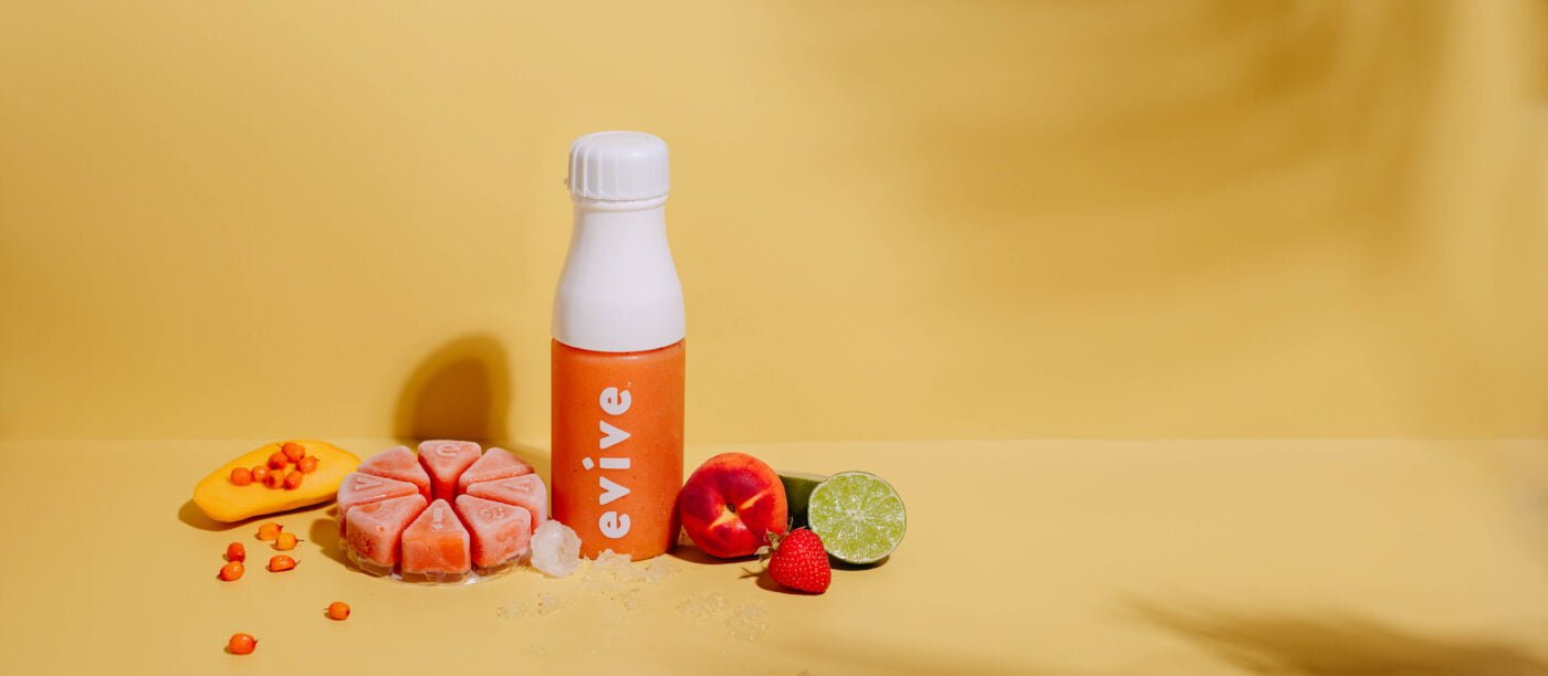 A bottle of Sunrize Smoothie made by Evive Smoothies. with some fruits and a smoothie wheel next to it. Sunrize smoothie has sea buckthorn berries, peaches, and strawberries.