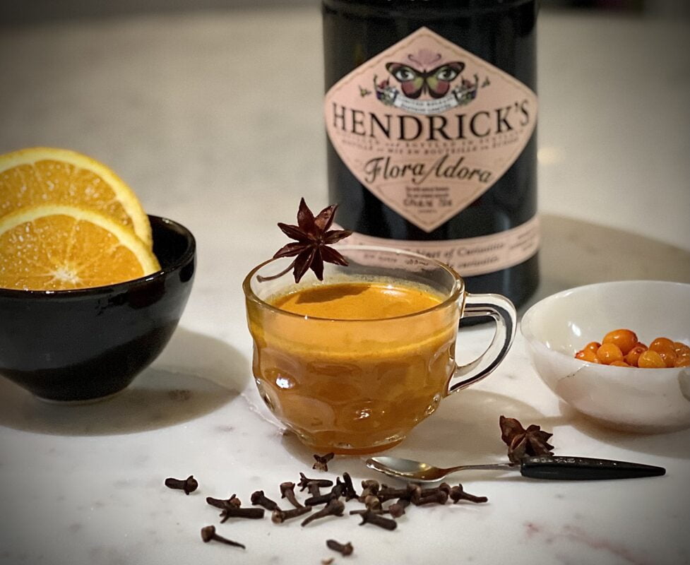 A Seaberry Hot Toddy in a glass with a bottle of Hendrix Flora Adora Gin and ingredients as decorations surrounding the glass.