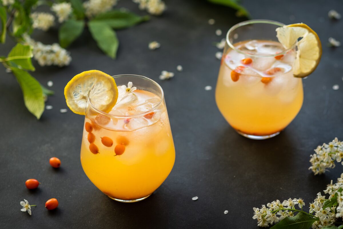 Two fresh cocktails with sea buckthorn, juice, and ice in glasses on dark background with seaberries and wild flowers at corners.